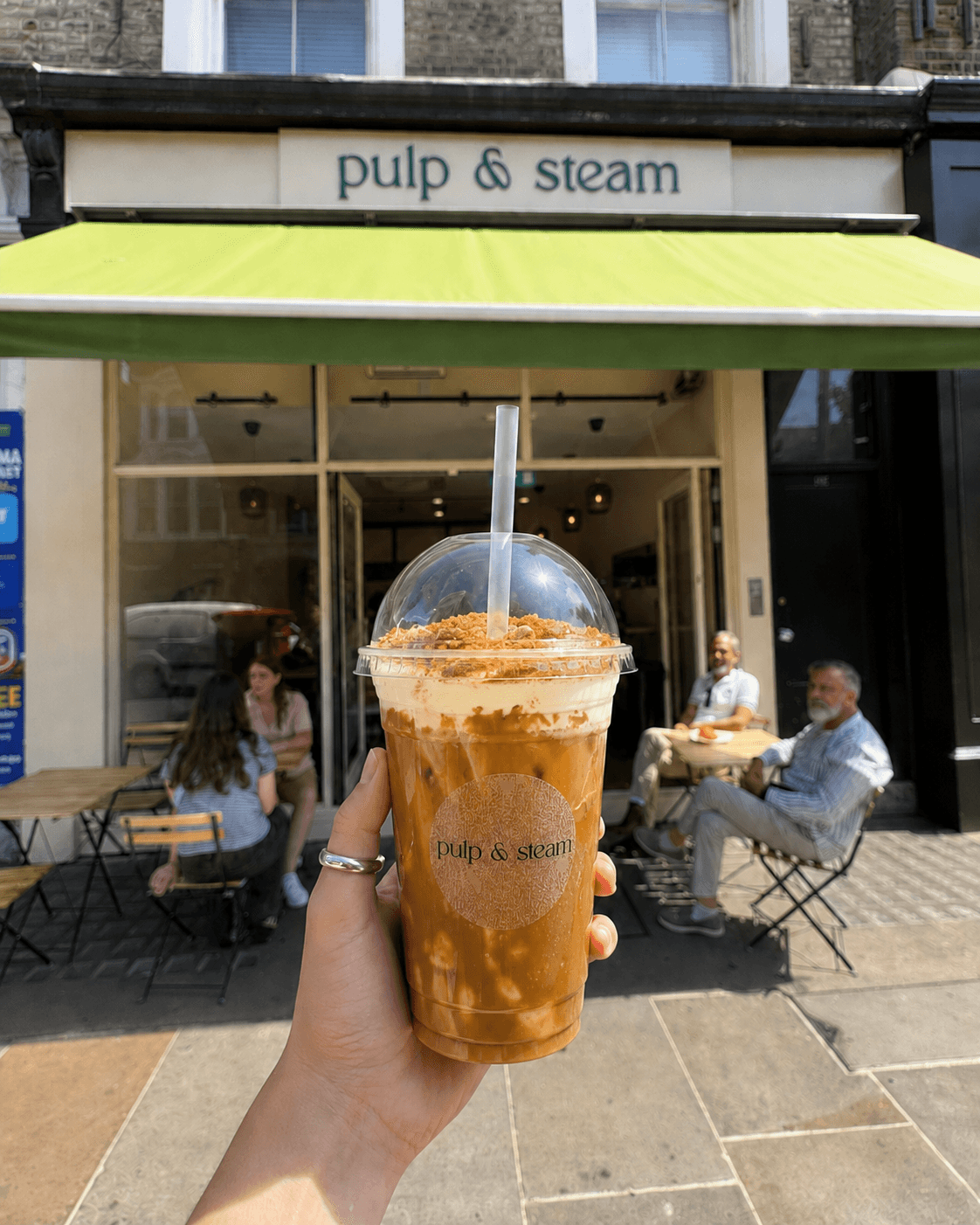 An iced coffee held in front of Pulp & Steam, with people sitting beneath the green awning on Ladbroke Grove